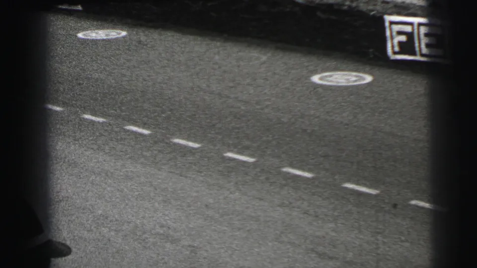 1954 Le Mans frame with the race surface, dashed lane markings, circular symbols and a dark blurred passing vehicle at the edge.