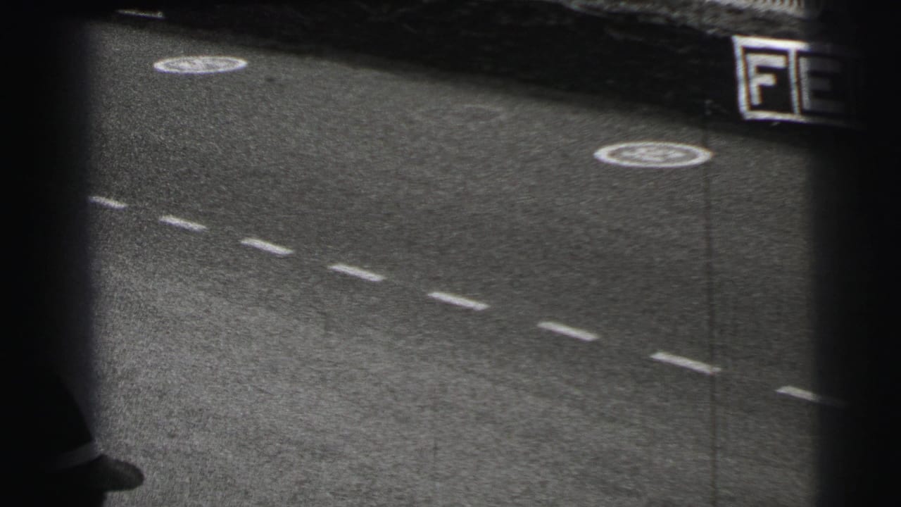 Le Mans, France frame from 1954 showing a grainy racetrack surface and a blurred vehicle edge moving out of view.