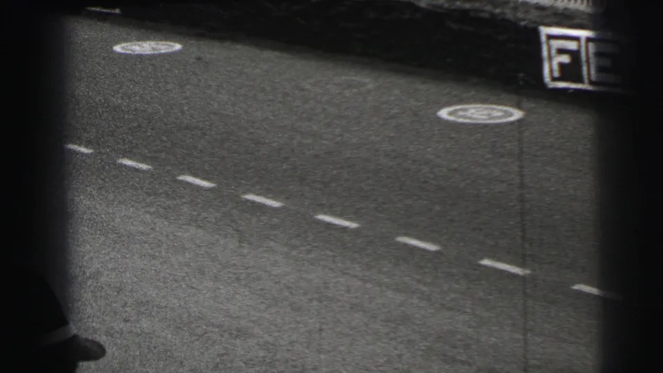 Le Mans, France frame from 1954 showing a grainy racetrack surface and a blurred vehicle edge moving out of view.