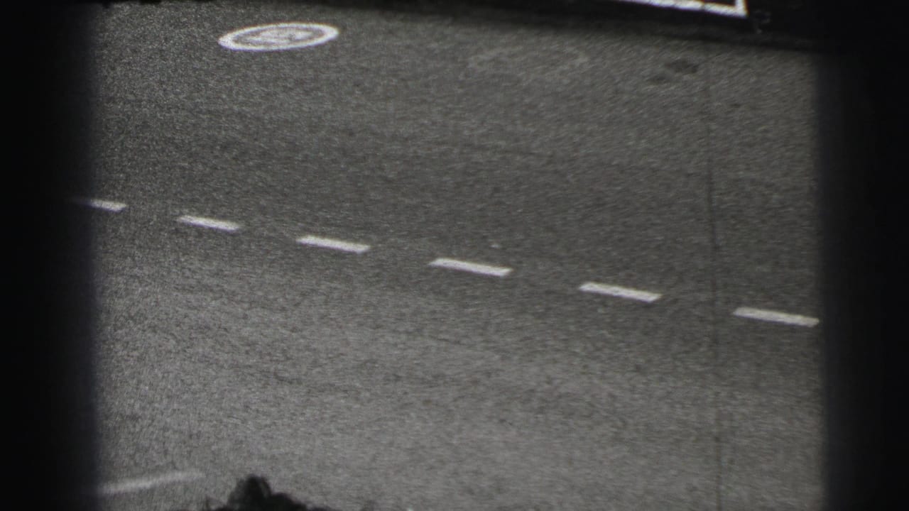 Black-and-white 8mm frame of the Le Mans track surface with painted lane markings and heavy film-edge shadow.