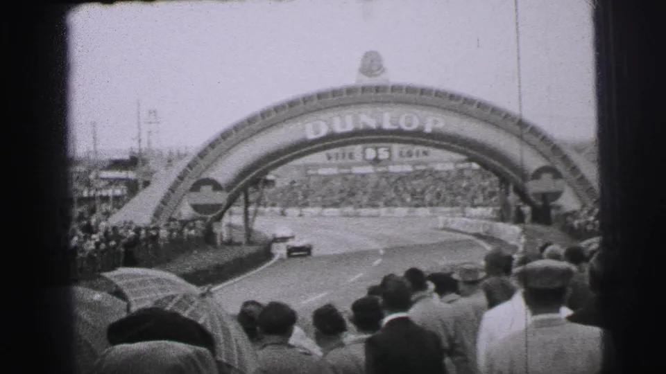 Le Mans race car passes under the Dunlop arch before a dense crowd of spectators in 1950s France.