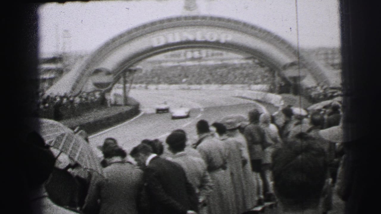 Two cars run toward the Dunlop arch at Le Mans as spectators with umbrellas watch from the foreground.