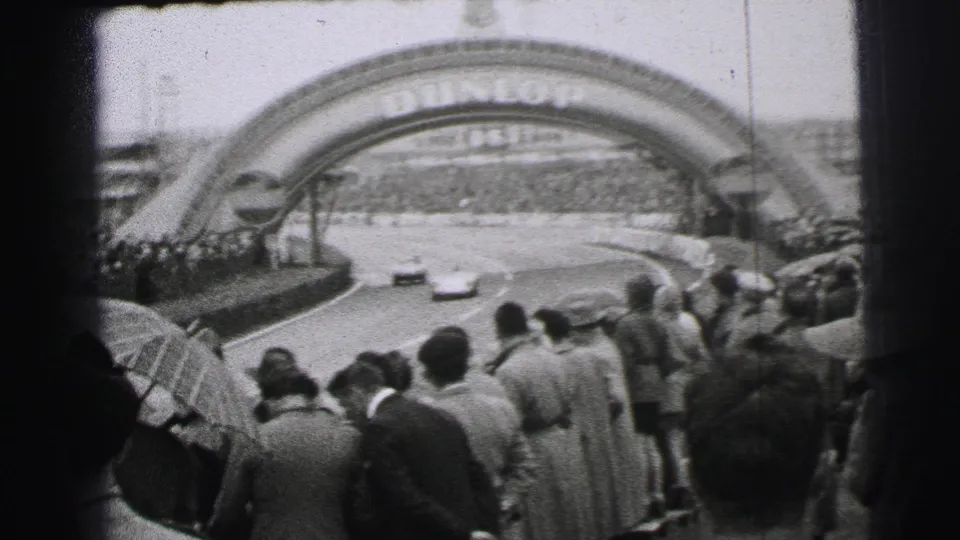 Two cars run toward the Dunlop arch at Le Mans as spectators with umbrellas watch from the foreground.