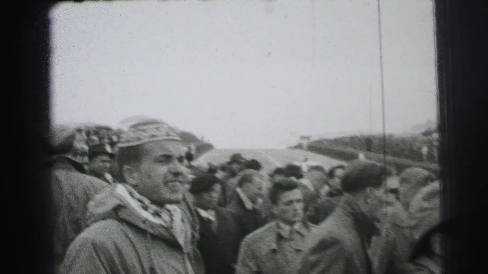 1954 Le Mans crowd view with a smiling man in a decorated hat and scarf standing among other spectators near the circuit.