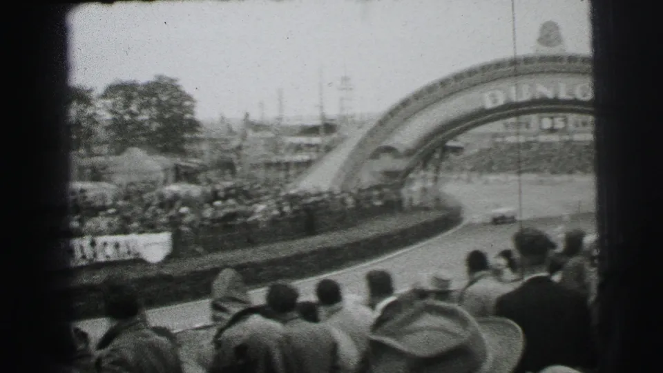 1954 Le Mans 8mm frame with cars on the circuit below the Dunlop bridge and a dense spectator crowd in the foreground.