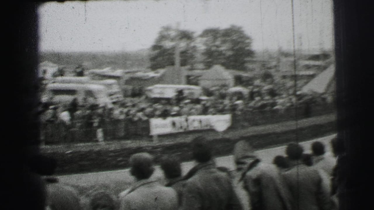 Crowd watches a race track from behind a barrier with cars and tents in the background.