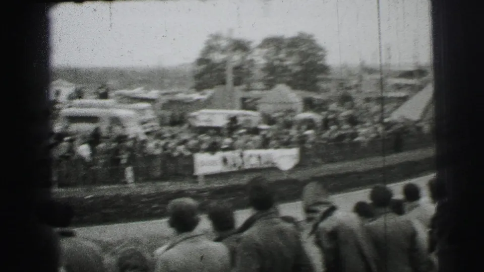 Spectators at Le Mans in 1954 appear along a trackside barrier, with parked vehicles, tents, and blurred trees beyond the course.