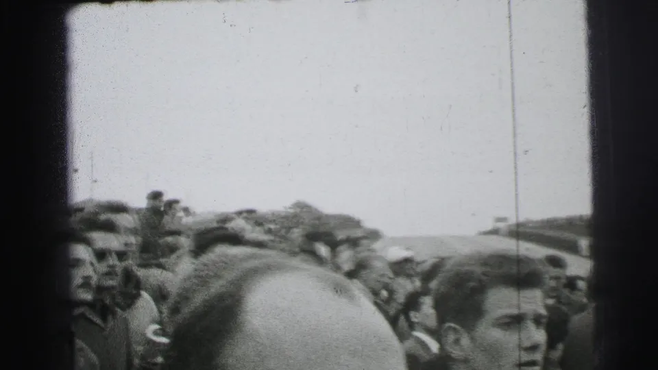 Le Mans spectators in 1950s clothing crowd near the course, with faces and heads filling the foreground of the handheld frame.