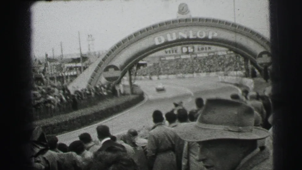 The Dunlop arch spans a curving section of the Le Mans circuit, with spectators lining the barriers and a car visible on the track.