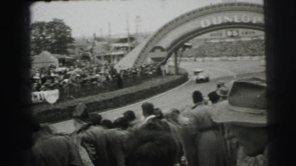 1954 Le Mans spectator-shot frame showing a racing car entering a curve under a Dunlop arch, with a dense crowd in the foreground.