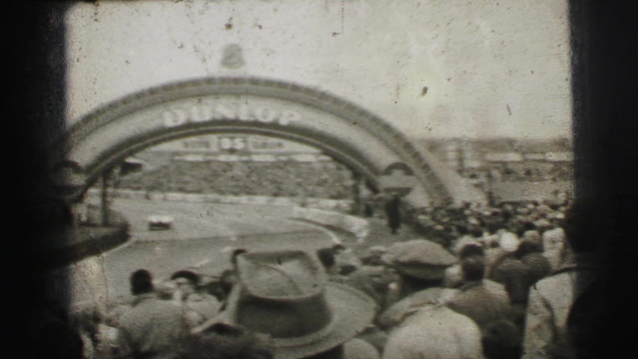 Le Mans home-movie frame showing a car rounding the course beneath the Dunlop arch while spectators fill the foreground and far side.