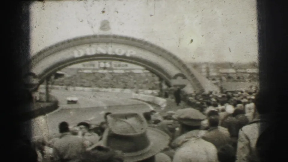 Le Mans home-movie frame showing a car rounding the course beneath the Dunlop arch while spectators fill the foreground and far side.