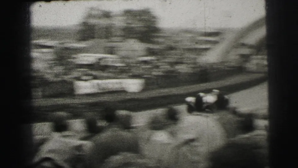 Handheld 1954 Le Mans frame with a blurred racing car, trackside spectators, barriers, and part of the Dunlop arch at the edge.