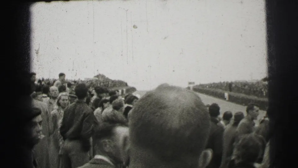 Crowd members in mid-century dress watch the Le Mans circuit from a trackside vantage point in 1954.