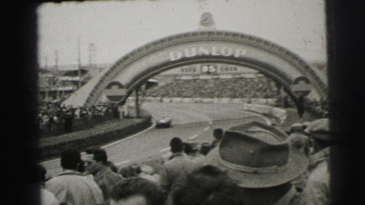 A race car moves under the Dunlop arch at Le Mans, with spectators and hats visible in the foreground.