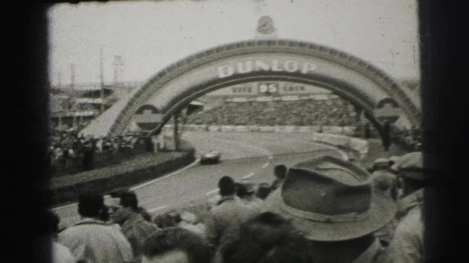 A race car moves under the Dunlop arch at Le Mans, with spectators and hats visible in the foreground.