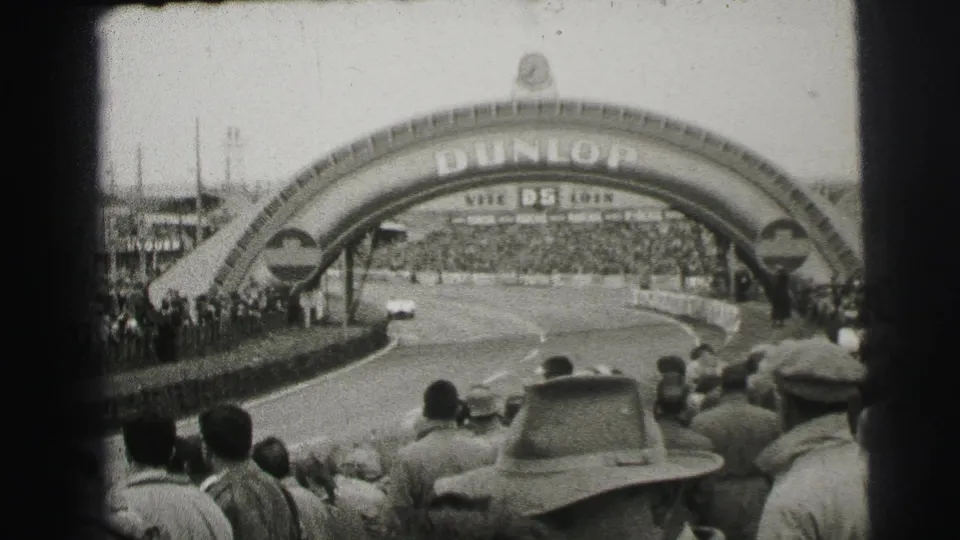 Crowds line the Le Mans circuit as race cars approach the Dunlop arch, with grandstands and track barriers visible in black-and-white 8mm footage.