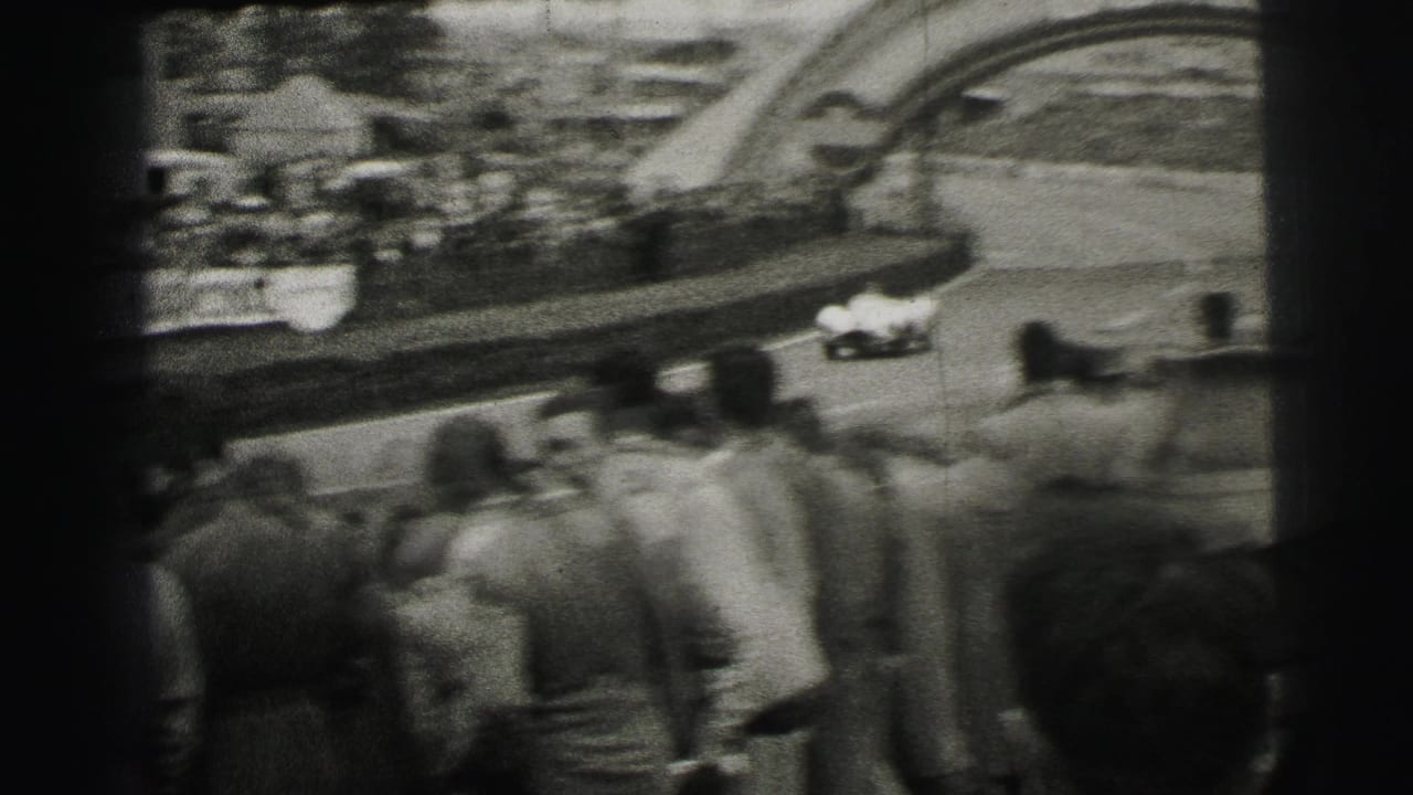 A white race car passes a crowd near the Dunlop arch at Le Mans, seen from a spectator position in grainy black-and-white 8mm footage.