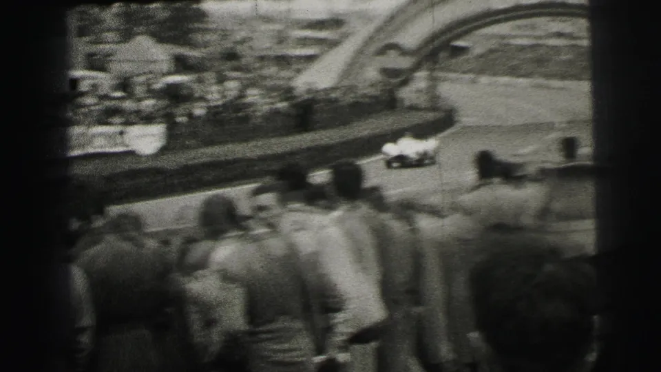 A white race car passes a crowd near the Dunlop arch at Le Mans, seen from a spectator position in grainy black-and-white 8mm footage.
