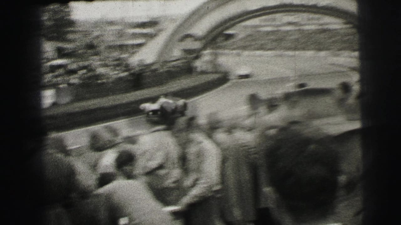From within the crowd, the camera frames race cars on the Le Mans circuit beneath the Dunlop arch, with spectators and track fencing in view.