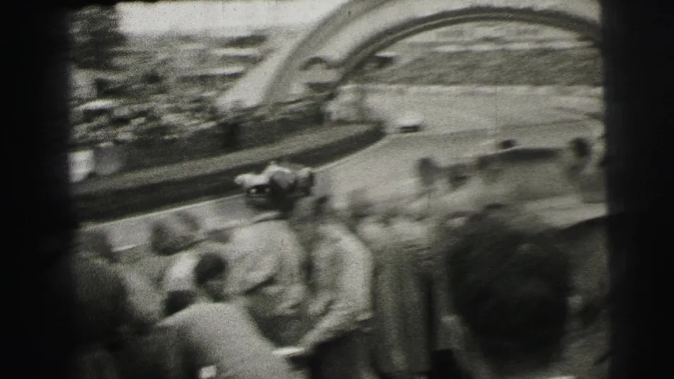 From within the crowd, the camera frames race cars on the Le Mans circuit beneath the Dunlop arch, with spectators and track fencing in view.
