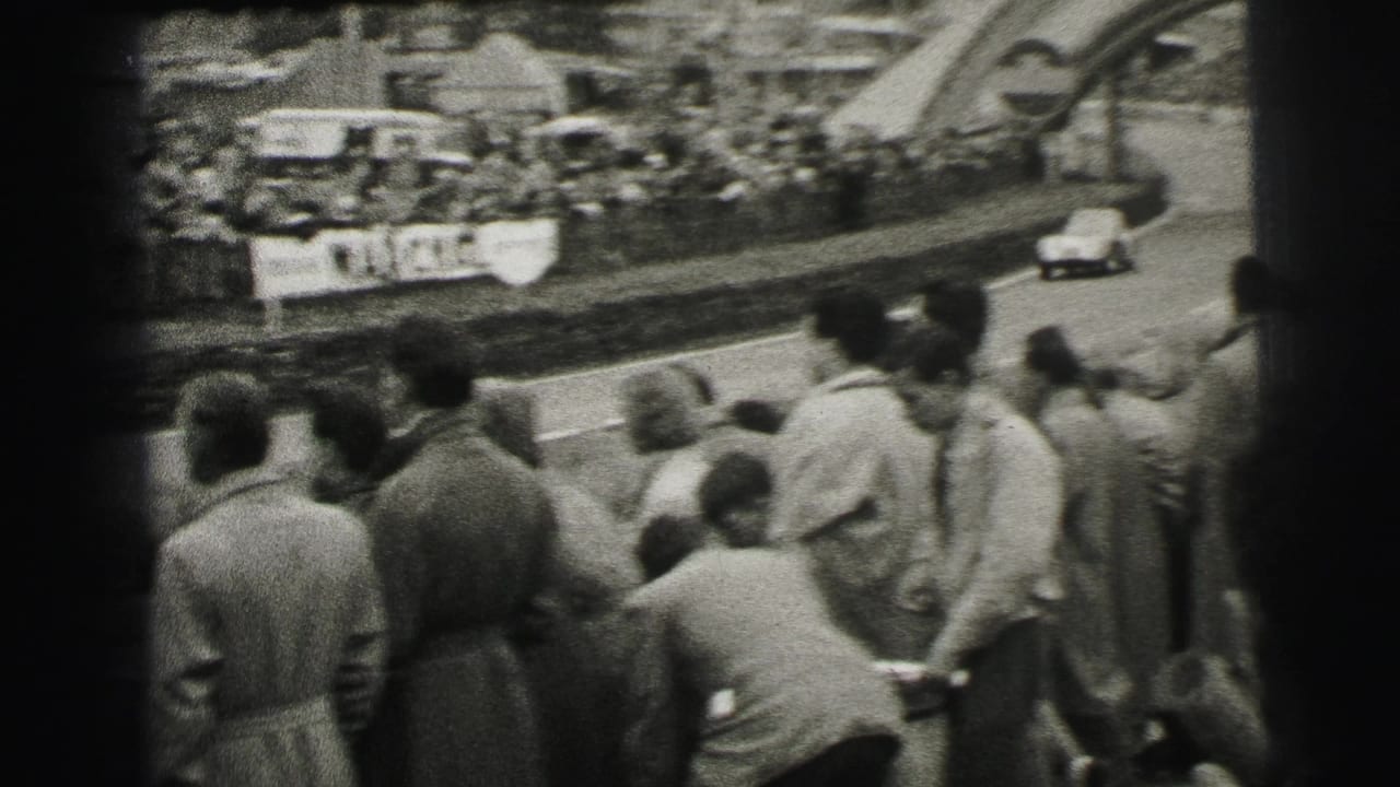 Spectators stand beside a race track as a small car approaches near banners.