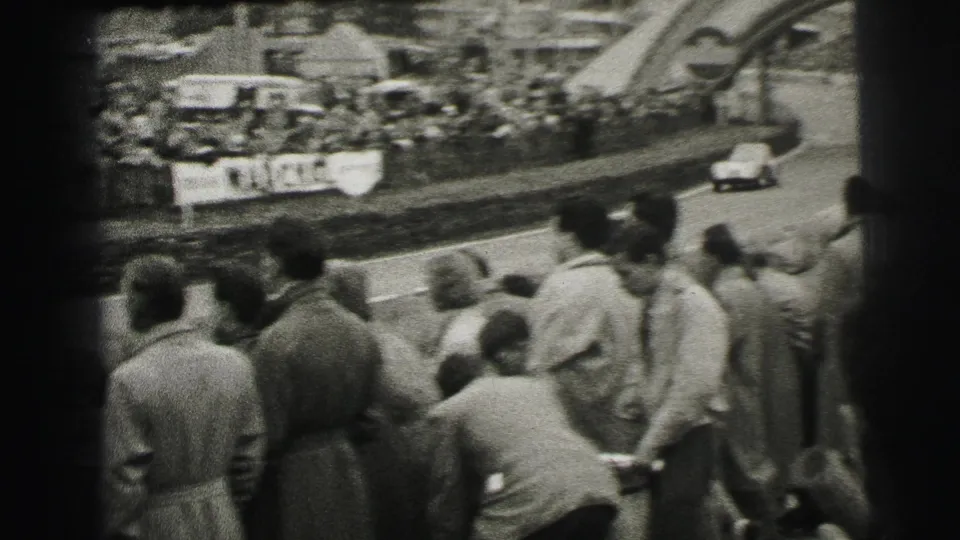 1954 Le Mans home-movie frame showing trackside spectators watching a racing car pass near banners and the course edge.