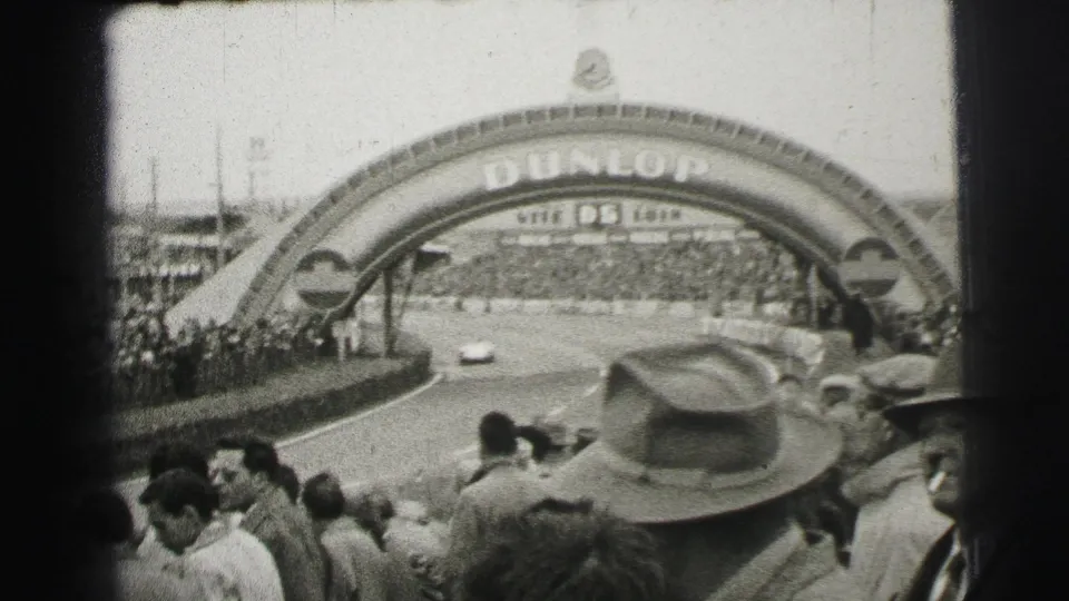 1954 Le Mans frame with a racing car under the Dunlop arch and crowded spectator areas lining the track.