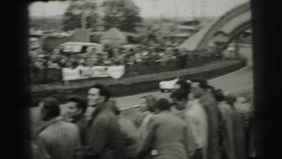 1954 Le Mans crowd view with spectators in mid-century dress watching a small racing car near the Dunlop arch area.