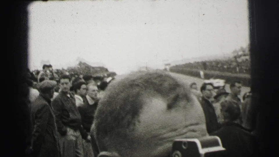1954 Le Mans spectator view with men gathered along the track and a large foreground head partially obscuring the race course.