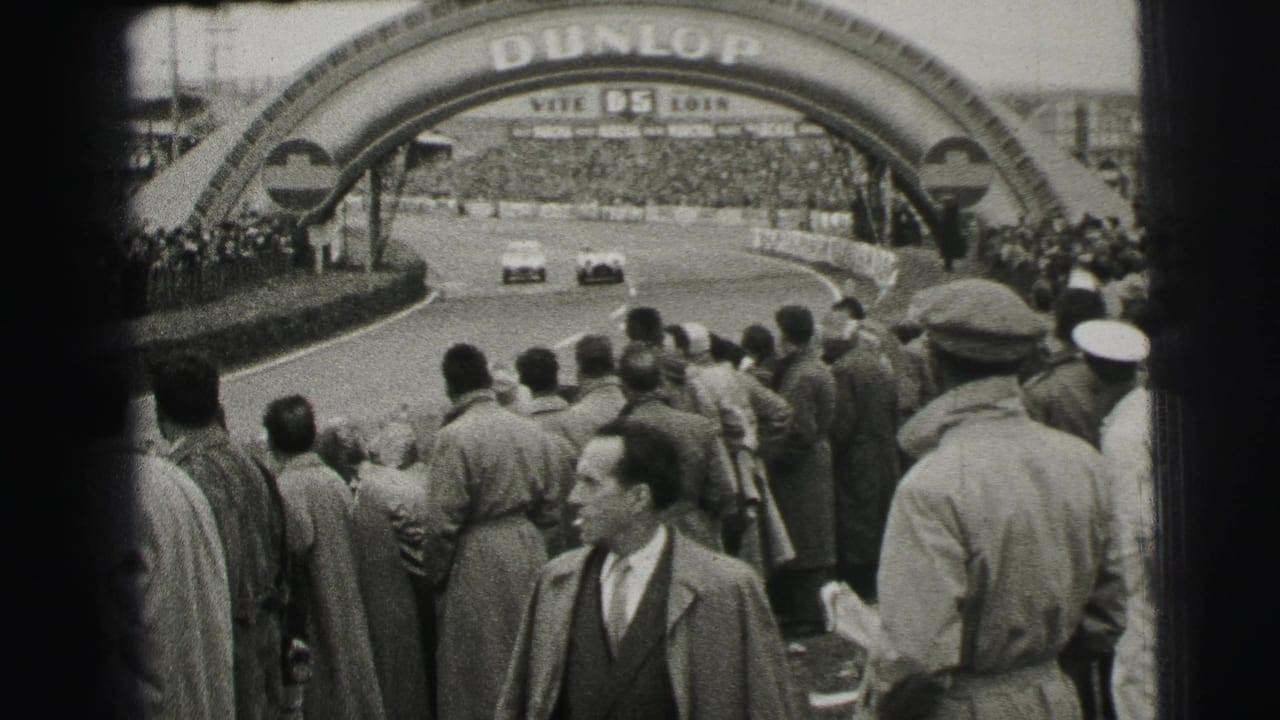 Two cars round the Le Mans circuit beneath the Dunlop bridge while a dense crowd of suited spectators watches from the edge.