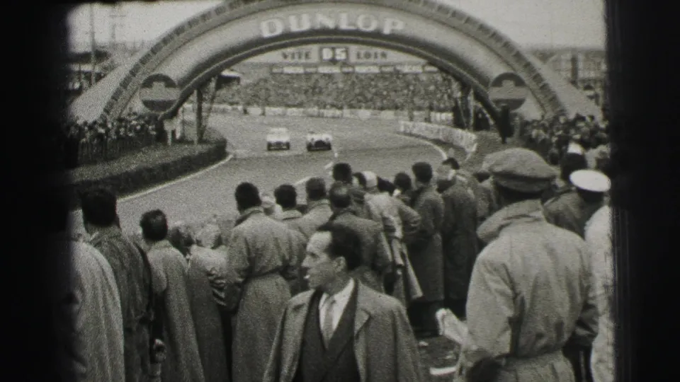 Two cars round the Le Mans circuit beneath the Dunlop bridge while a dense crowd of suited spectators watches from the edge.