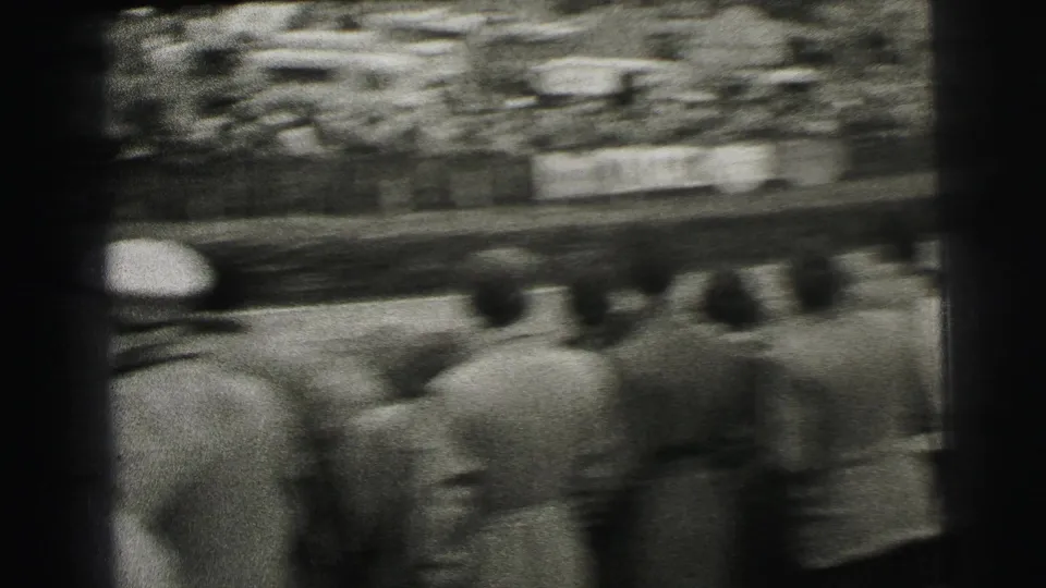 A race car streaks past the Le Mans crowd, with motion blur and spectators seen from behind near the circuit edge.