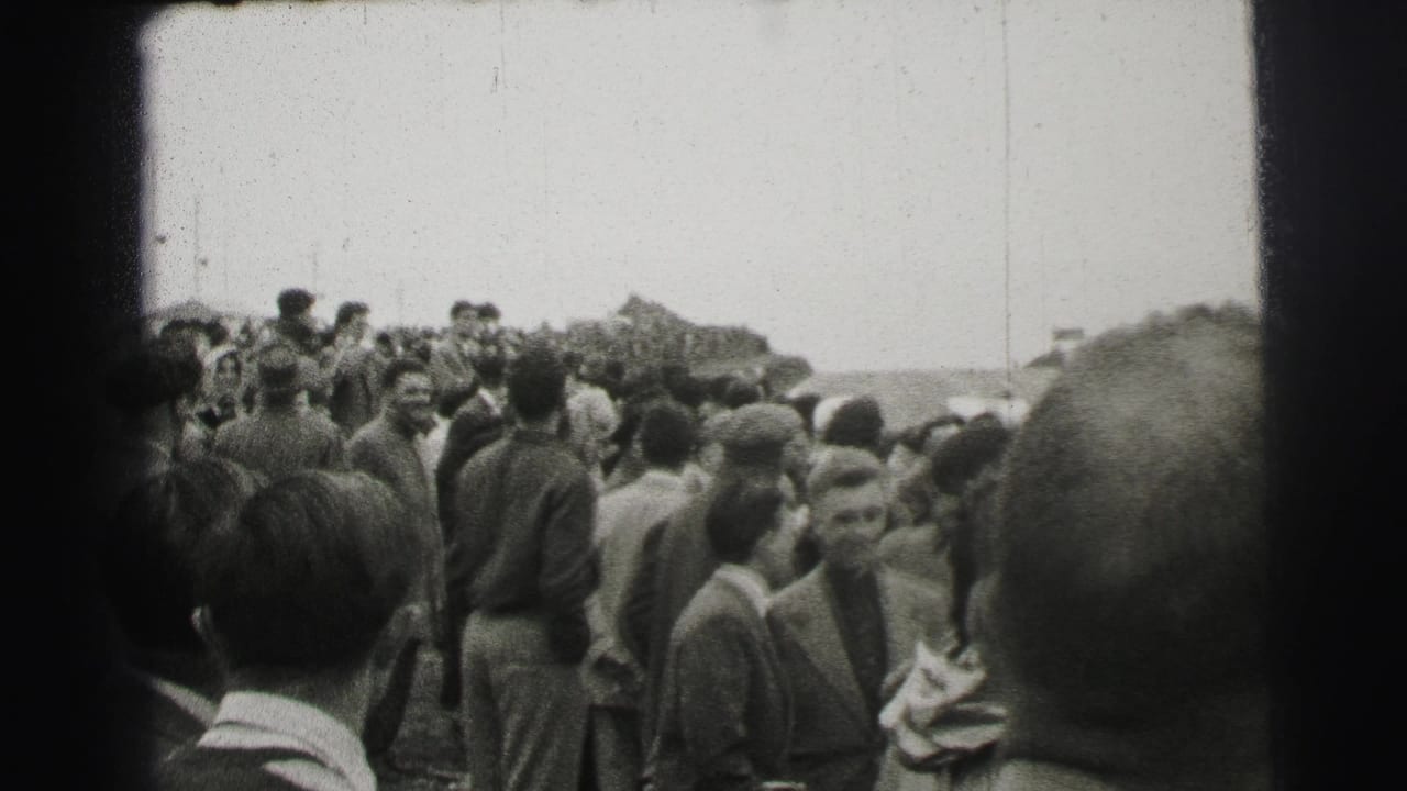 Crowd of standing spectators gathered beside a race course under an overcast sky.