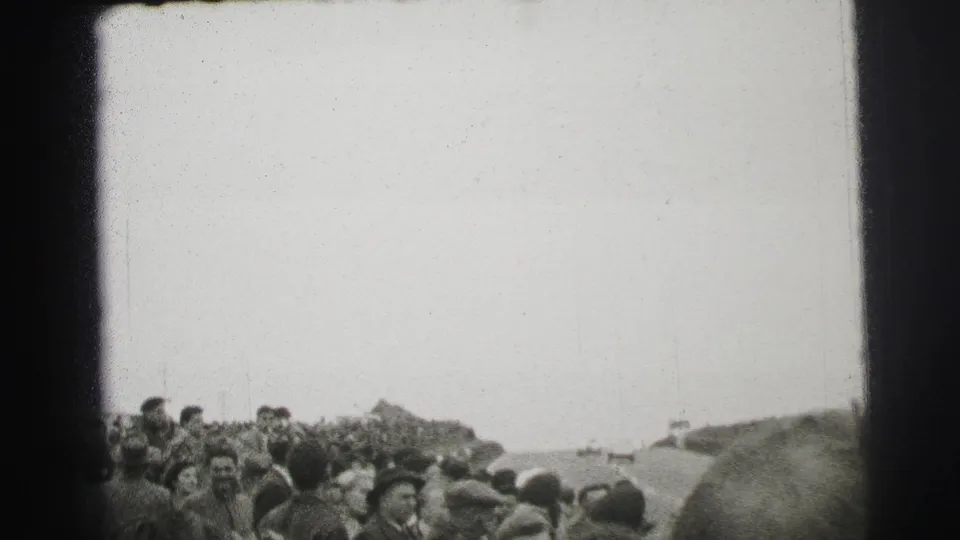 A dense Le Mans crowd watches from beside the course, with foreground heads and distant spectators visible in the 8mm frame.