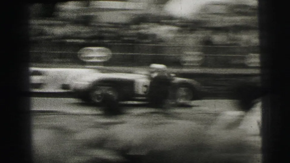 A race car blurs through the frame at Le Mans, emphasizing speed as spectators watch from behind the course edge.
