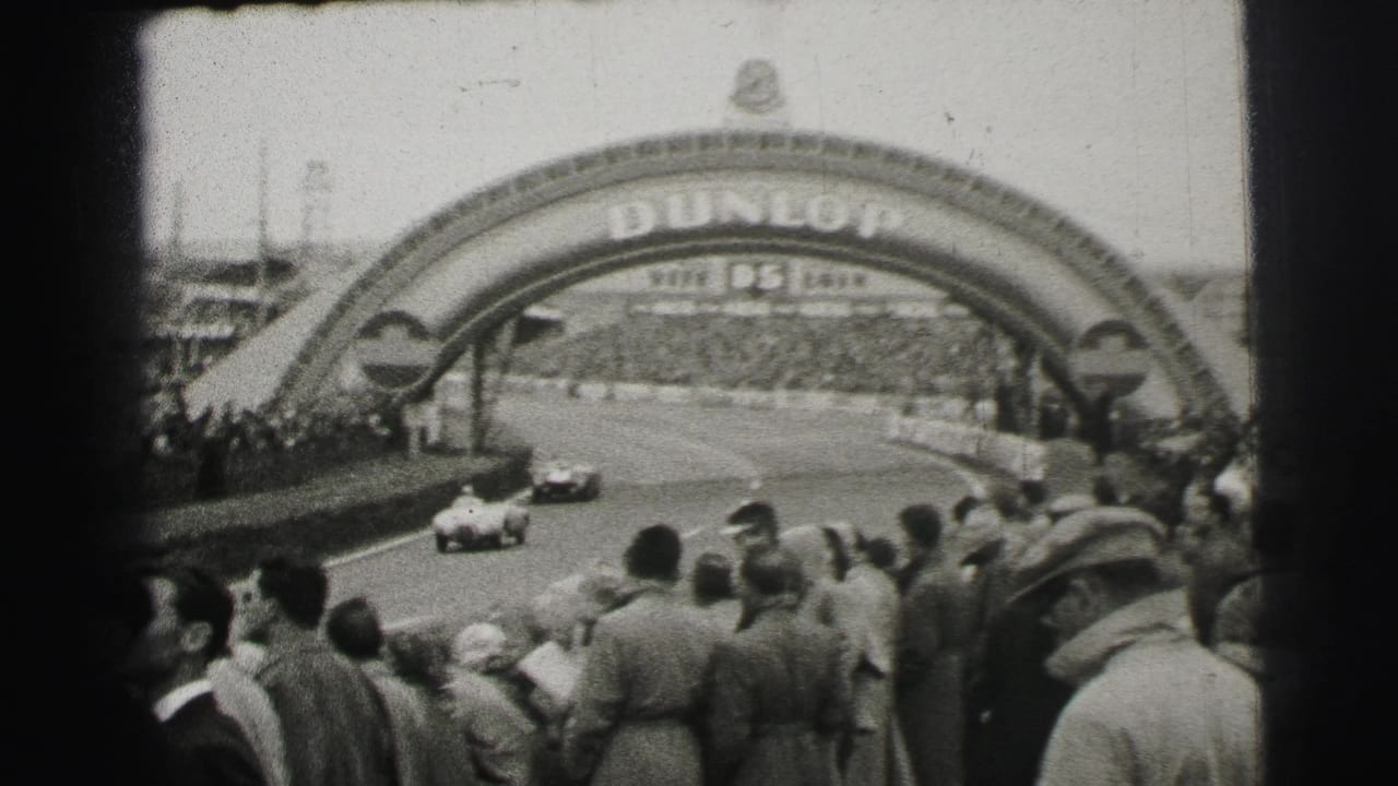 Race cars travel under the Dunlop bridge at Le Mans while a dense crowd watches from the side.