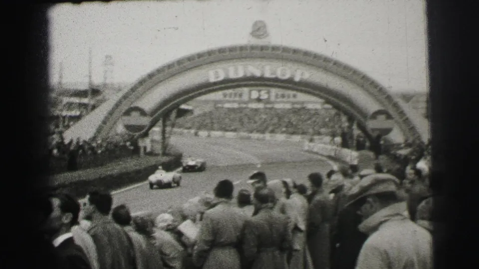 Race cars travel under the Dunlop bridge at Le Mans while a dense crowd watches from the side.