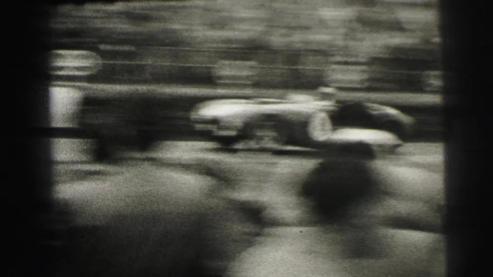 A low race car blurs across the frame with a packed Le Mans spectator area behind it.