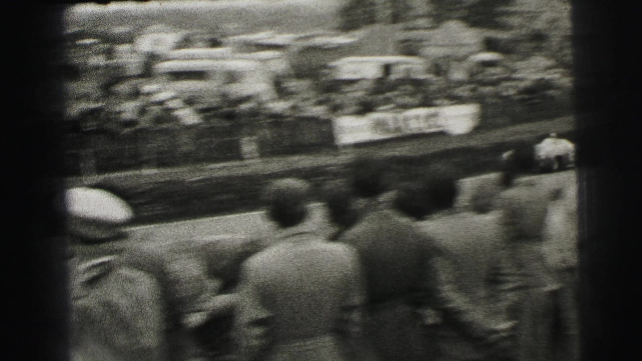 A Le Mans crowd viewed from behind as a blurred race car moves past on the circuit in 1954 home-movie footage.