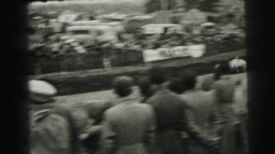 A Le Mans crowd viewed from behind as a blurred race car moves past on the circuit in 1954 home-movie footage.