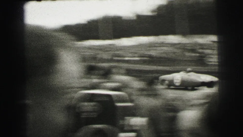Motion-blurred racing action at Le Mans, with cars and trackside spectators captured through handheld 8mm framing.
