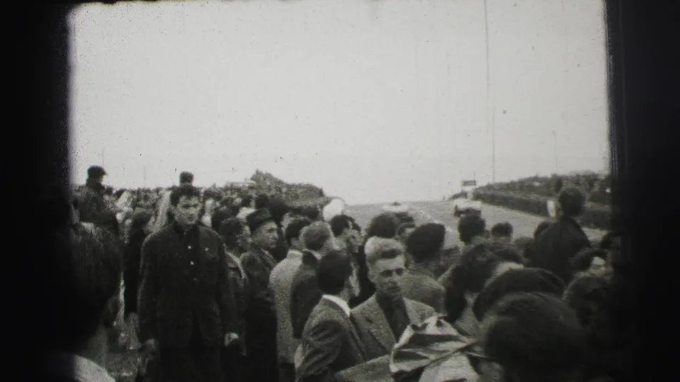 At Le Mans in 1954, spectators in coats gather close to the track while the handheld camera looks through the crowd.