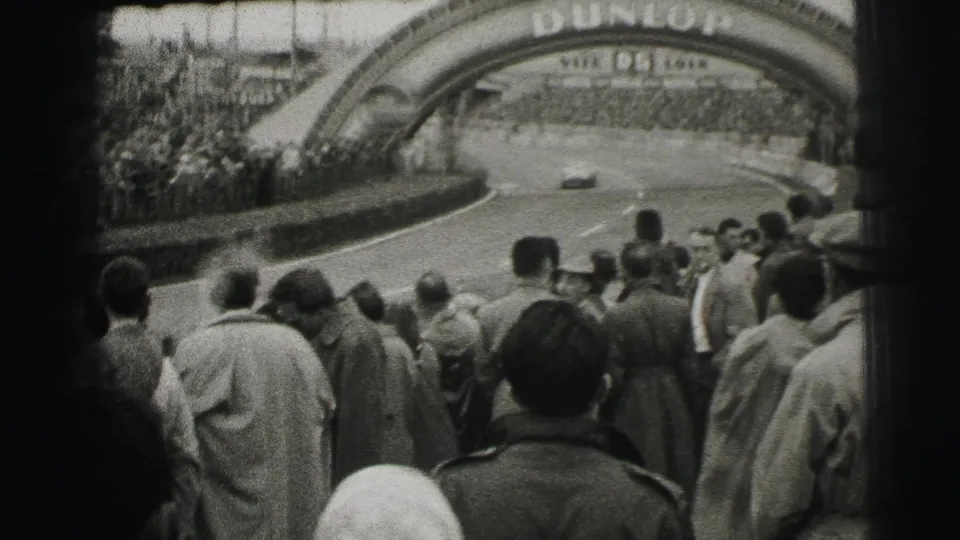 Spectators at the Le Mans circuit watch cars move through a bend beneath the visible Dunlop arch.