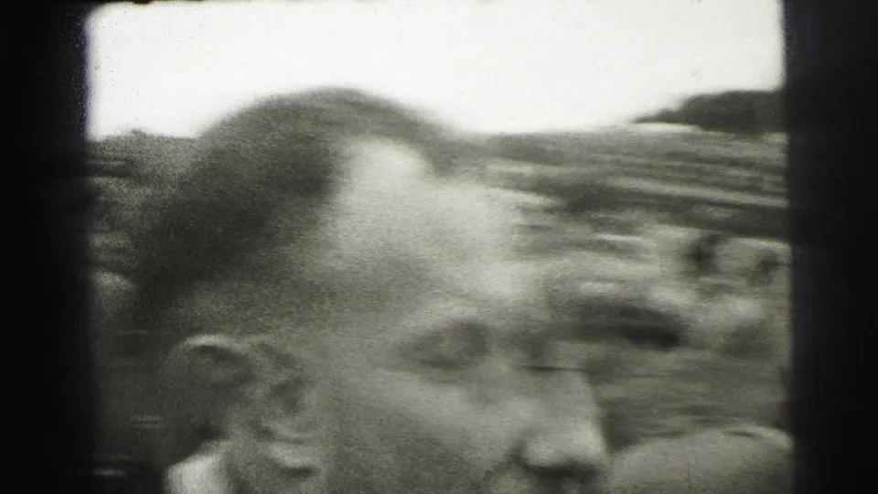 A close spectator profile fills the foreground as the Le Mans track and viewing crowd blur behind him.