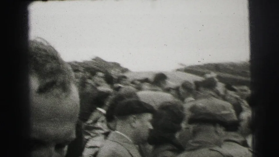 1954 Le Mans home-movie frame showing spectators in caps and coats near the track, partly obscured by a close foreground face.