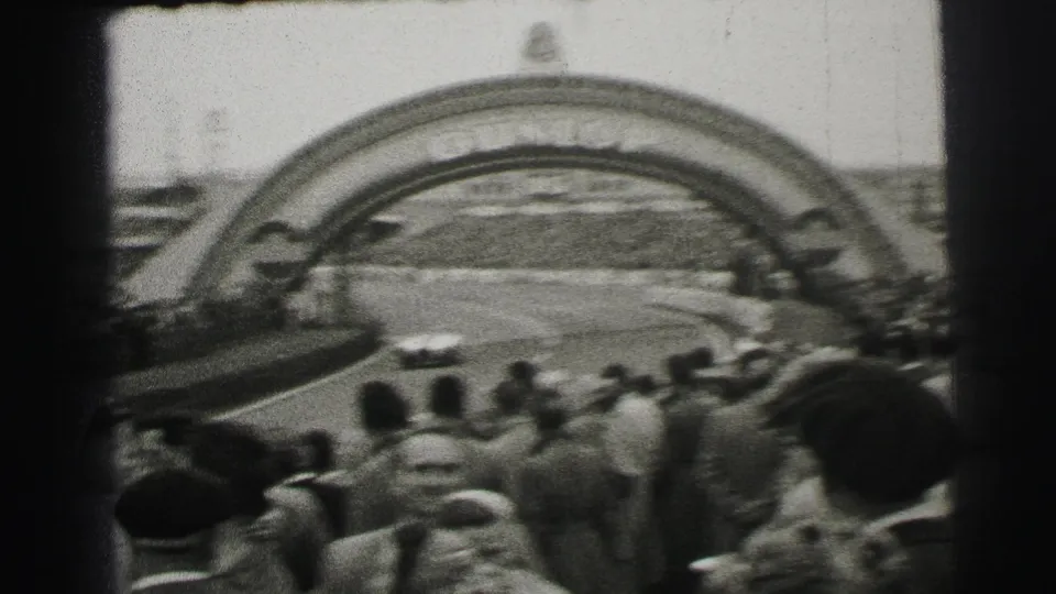Trackside crowd at Le Mans, France, looking toward the course with a large racing arch visible beyond the spectators.