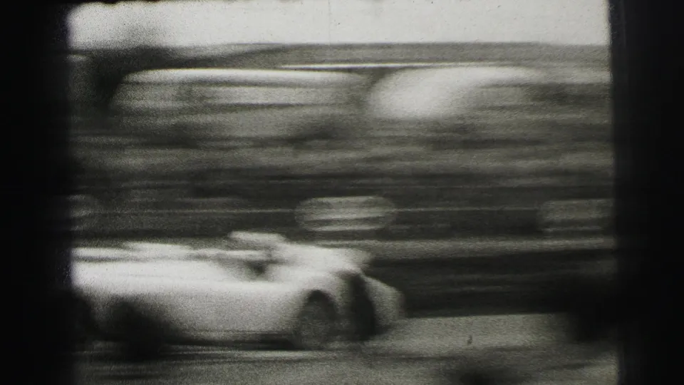 A fast race car blurs past the Le Mans crowd, emphasizing speed in grainy black-and-white 8mm footage.