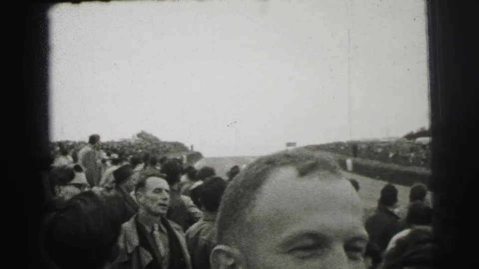 Le Mans spectators stand along the track as a foreground man's face and head partially obscure the home-movie view.