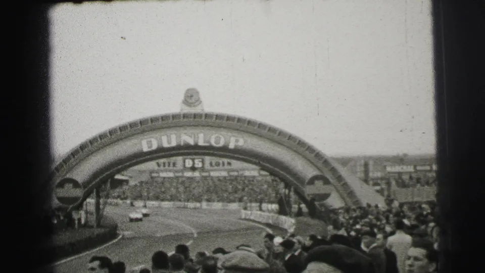 1954 Le Mans frame showing open race cars passing under the Dunlop arch as large crowds watch from the circuit edges.